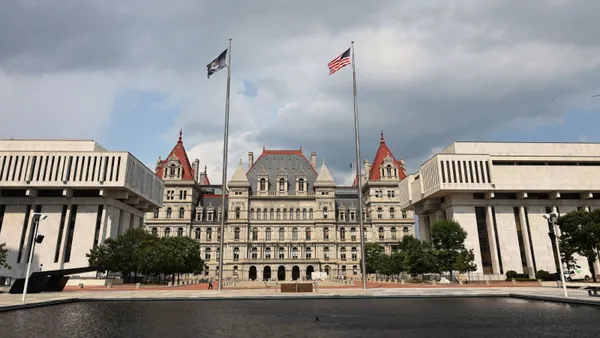 A large building with a ref roof is flanked by two square buildings in front of it. Two flags on flagpoles stand in the foreground.