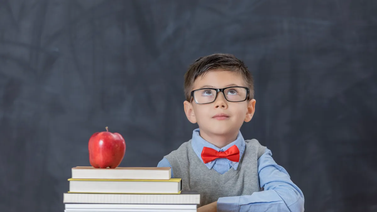 A young student sits at a desk in front of a blackboard. On the desk are a stack of books and a red apple. The student is wearing glasses and looking toward the sky.