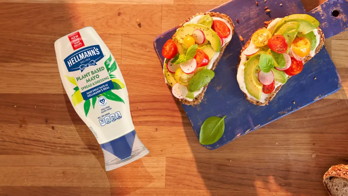 A bottle of plant-based Hellmann's mayo in a squeeze bottle next to an open-faced sandwich with vegetables on bread.