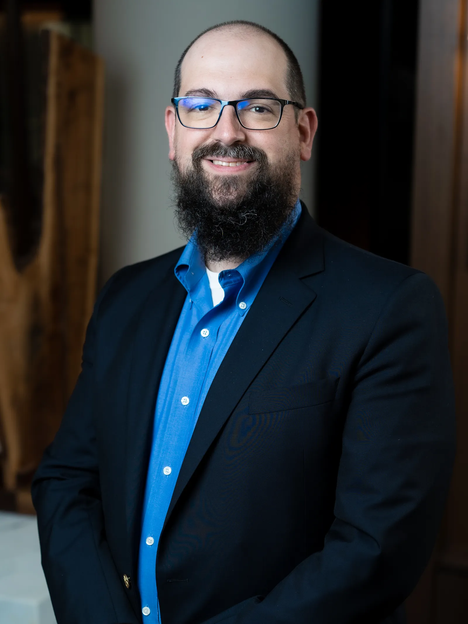 A portrait headshot of a man with a large dark beard and glasses