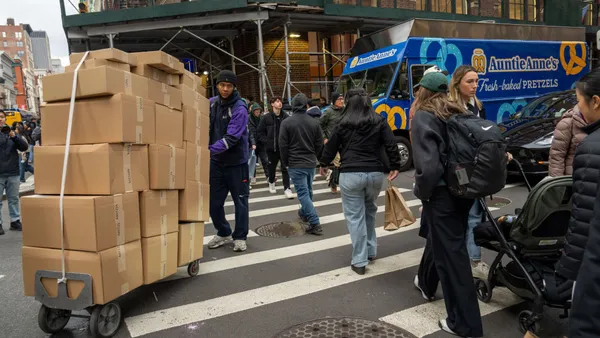 A person pushing a cart stacked with boxes navigates around people on a busy crosswalk.