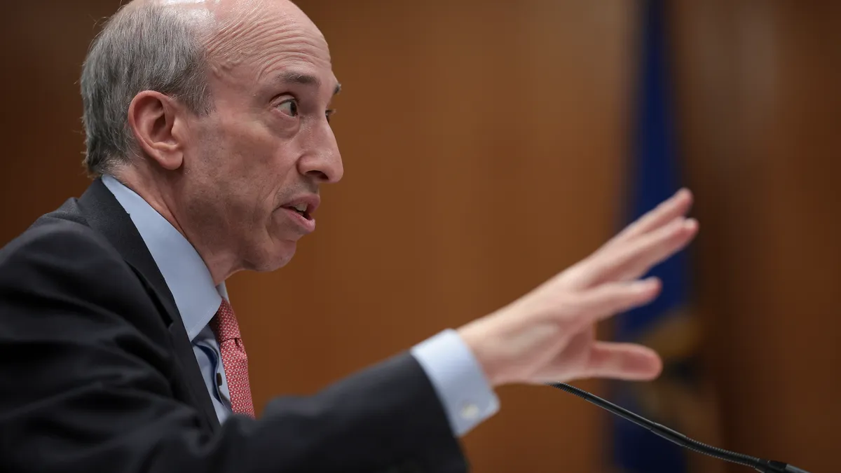 Gary Gensler speaks with his hand outstretched, seated before a microphone.