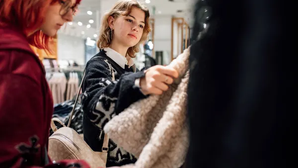 Two women browse a clothing rack in a bright boutique, selecting a plush beige coat.