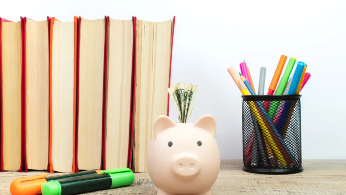 A piggy bank with a bill folded in the slot stands on a table next to a row of books, a cup of colorful pens and pencils and highlighters laying on the table.