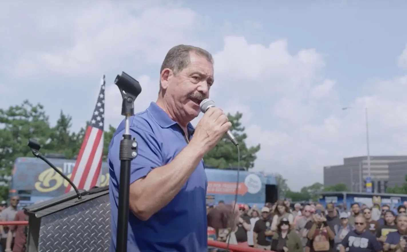 A person on an outdoor podium speaks into a microphone to a crowd of people.