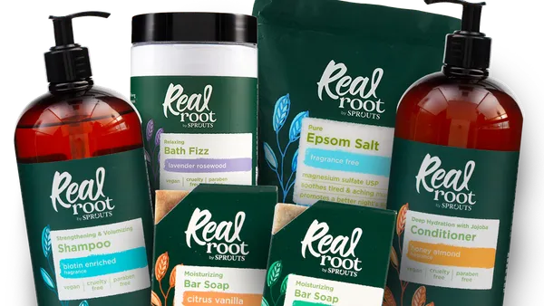 Array of Sprouts Real Root products, including bottles of shampoo and conditioner, bar soaps, bath fizz and Epsom salt all in dark green packaging against a dark gray background.