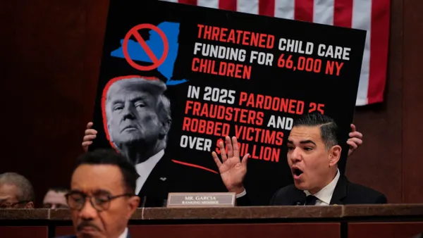 A person wearing a suit and tie with a placard on desk that says "Mr. Garcia" in front of a sign with picture of President Donald Trump that says "Threatened Child Care Funding for 66,000 NY Children. In 2025 Pardoned 25 Fraudsters and Robbed Victims"