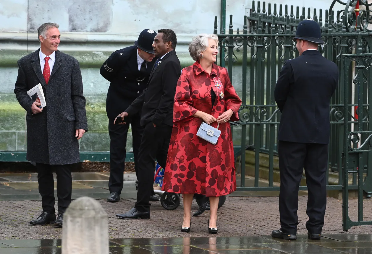 A woman in a red floral coat stands among a group of black-coated British guards, smiling.