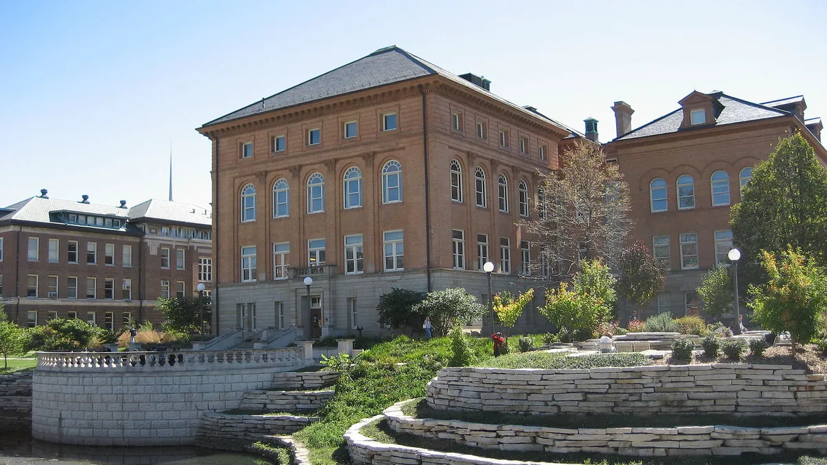 A tall brick building stands behind some green grass and the open sky. The building is old-seeming, with many windows.