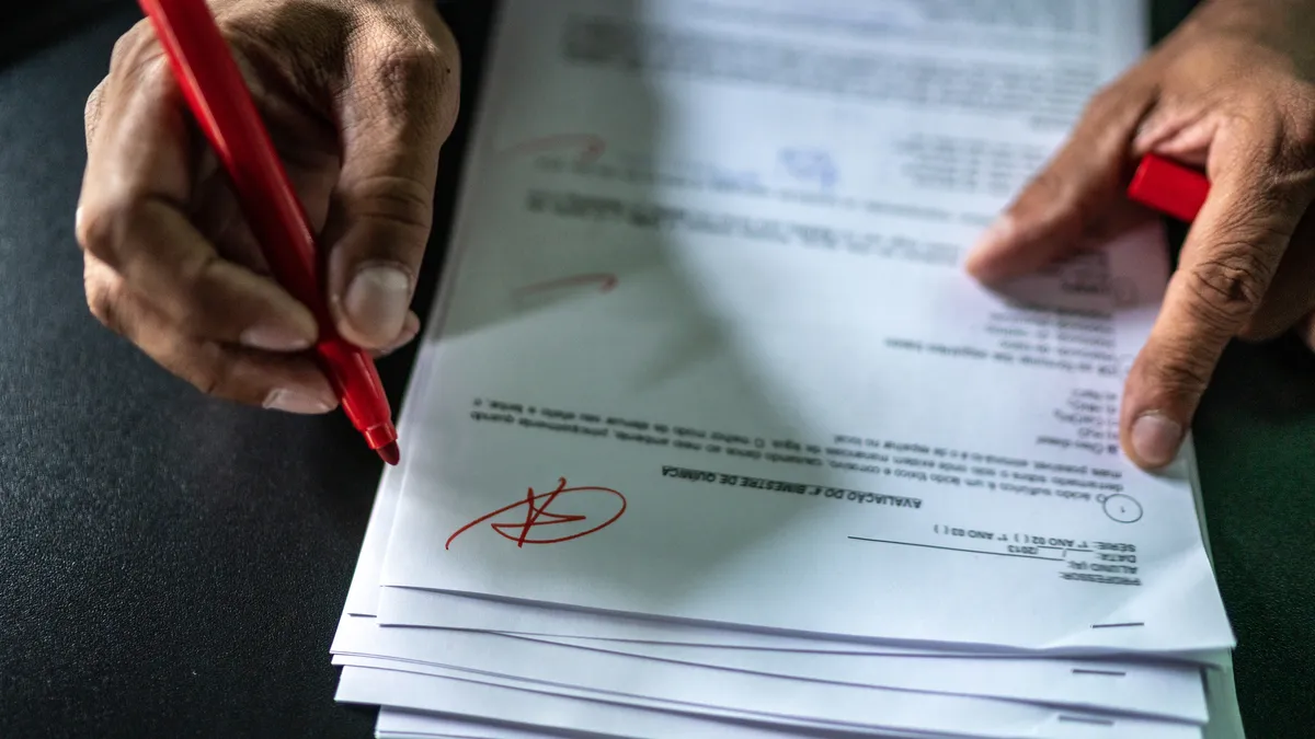 A closeup photo of a pair of hands. holding a stack of papers. In one hand is a red marker.