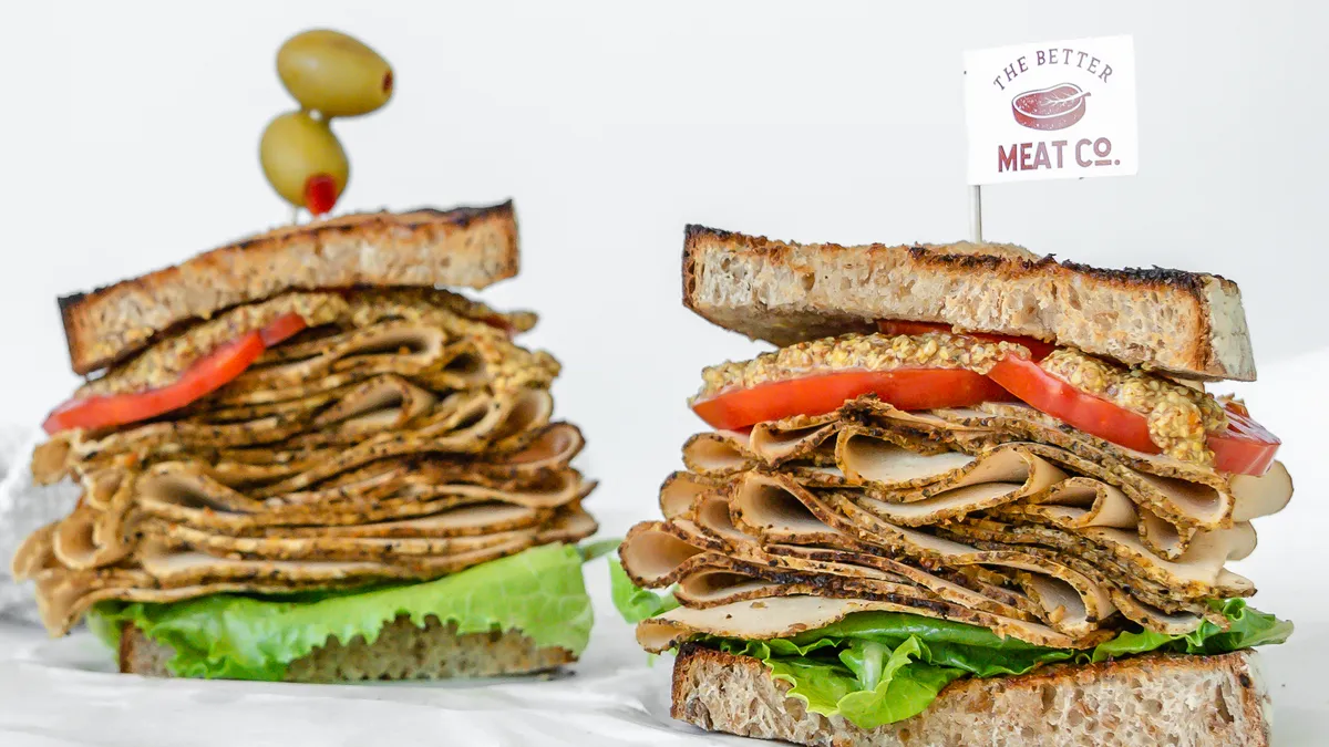 Two sandwich halves are stacked high with deli slices that look like turkey made from The Better Meat Co.'s Rhiza protein, with a company logo flag on a toothpick stuck in one.