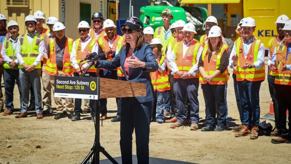 A person at a podium with the sign "Second Ave. Subway-Next stop 125th St." with people in construction uniforms behind.