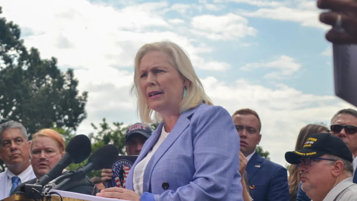 A blonde woman in a powder blue suit speaking into a mic, surrounded by people.