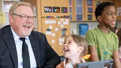 An adult sits at a desk. Two students are nearby by and everyone is smiling