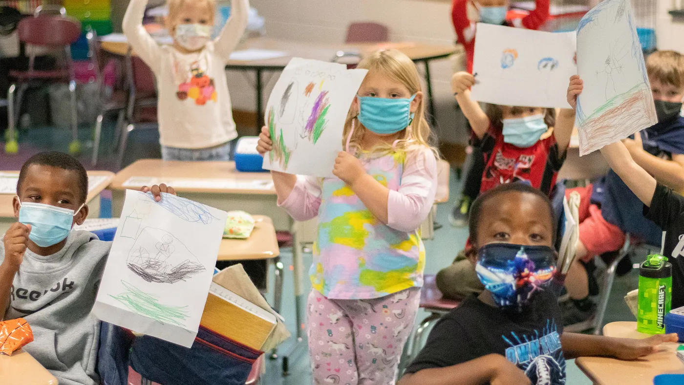 Elementary students in a classroom hold up their drawings.