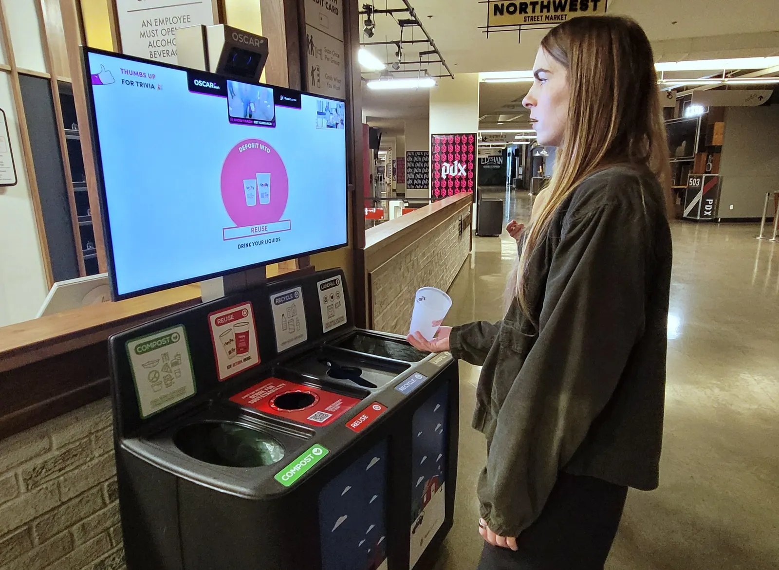 A person stands in front of a digital screen with a camera that is attached to a bin with sections for waste, recycling, reuse and composting.