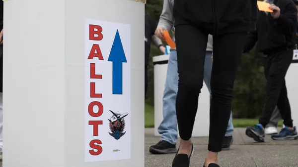 A person in black pants and shoes standing next to a light yellow box with "Ballot" in red text and a blue arrow pointing up.