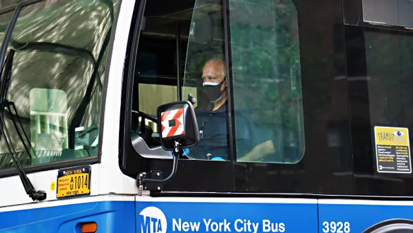 A bus driver wearing a mask seen through the driver's side window.