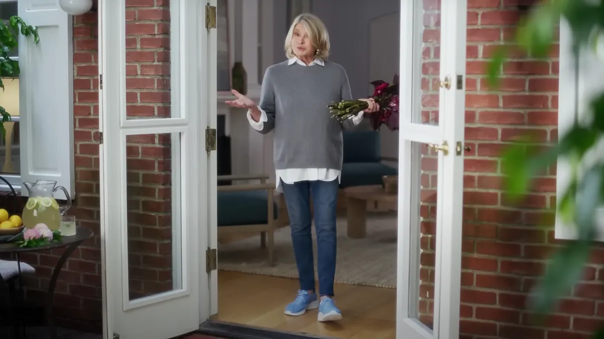 A person stands at a doorway wearing a sweater, jeans and sneakers while holding a bunch of cut flowers in one hand