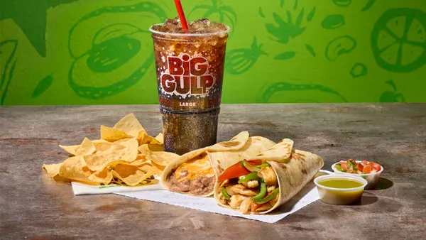 A meal consisting of a cup of soda, chips, salsa and tacos sits on a napkin on a wooden table against a green background. Text on the cup says Big Gulp Large.
