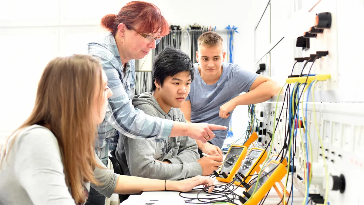 Three students are sitting in front of a panel with wires. An adult standing behind them is pointing to the panel