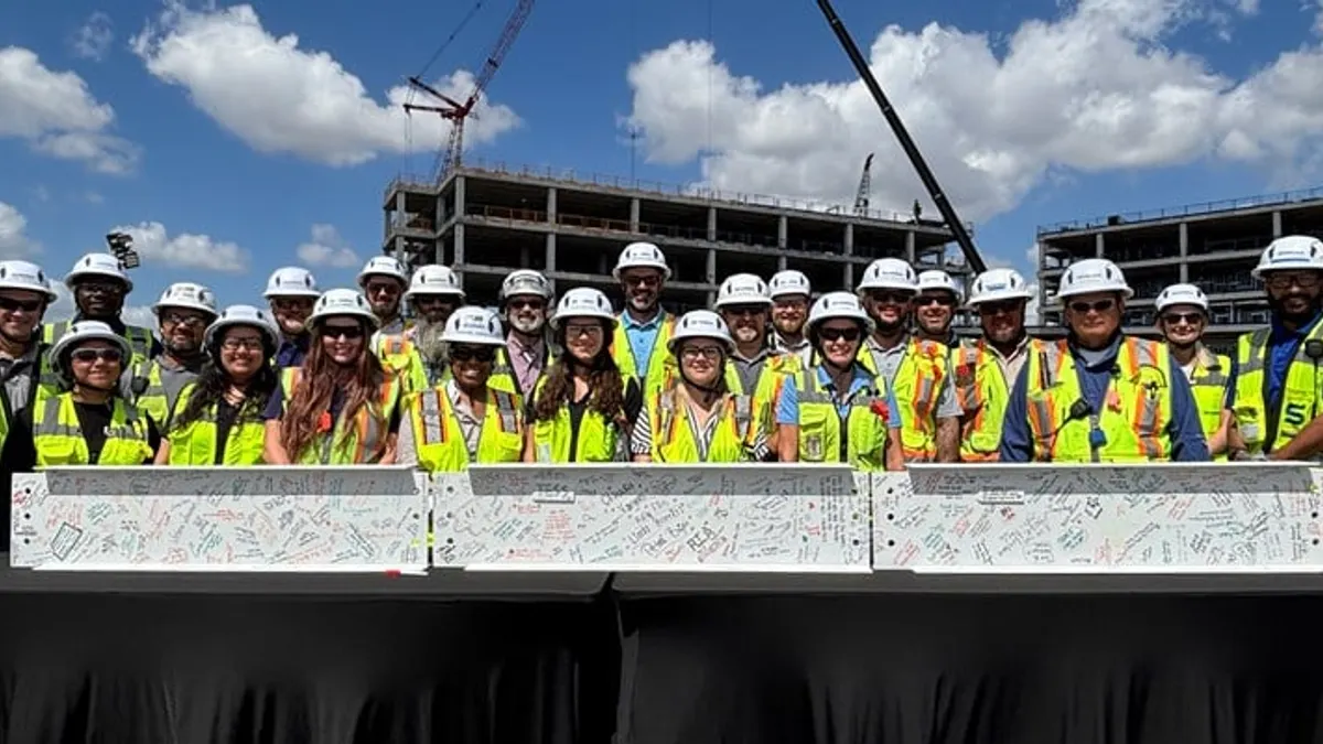 A group of people in vests and hard hats pose for a photograph behind a signed construction beam.