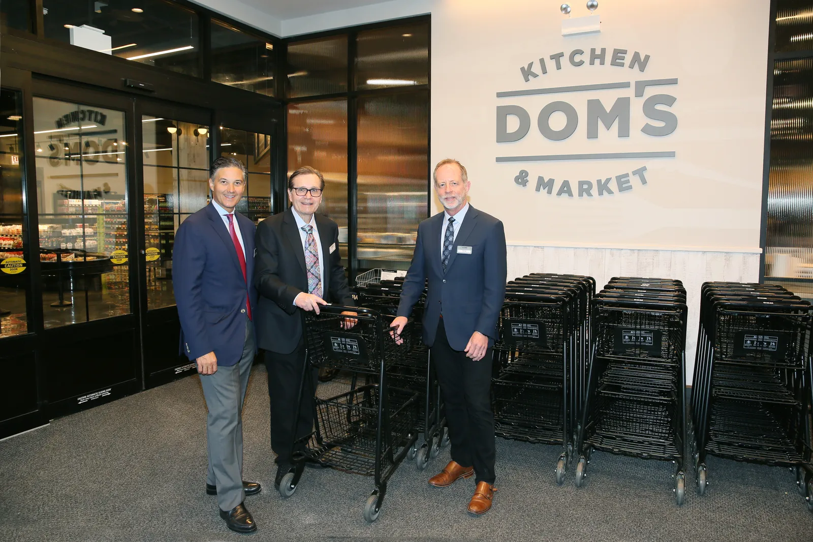 Three men standing in front of shopping carts and a sign that says Dom's Kitchen & Market.