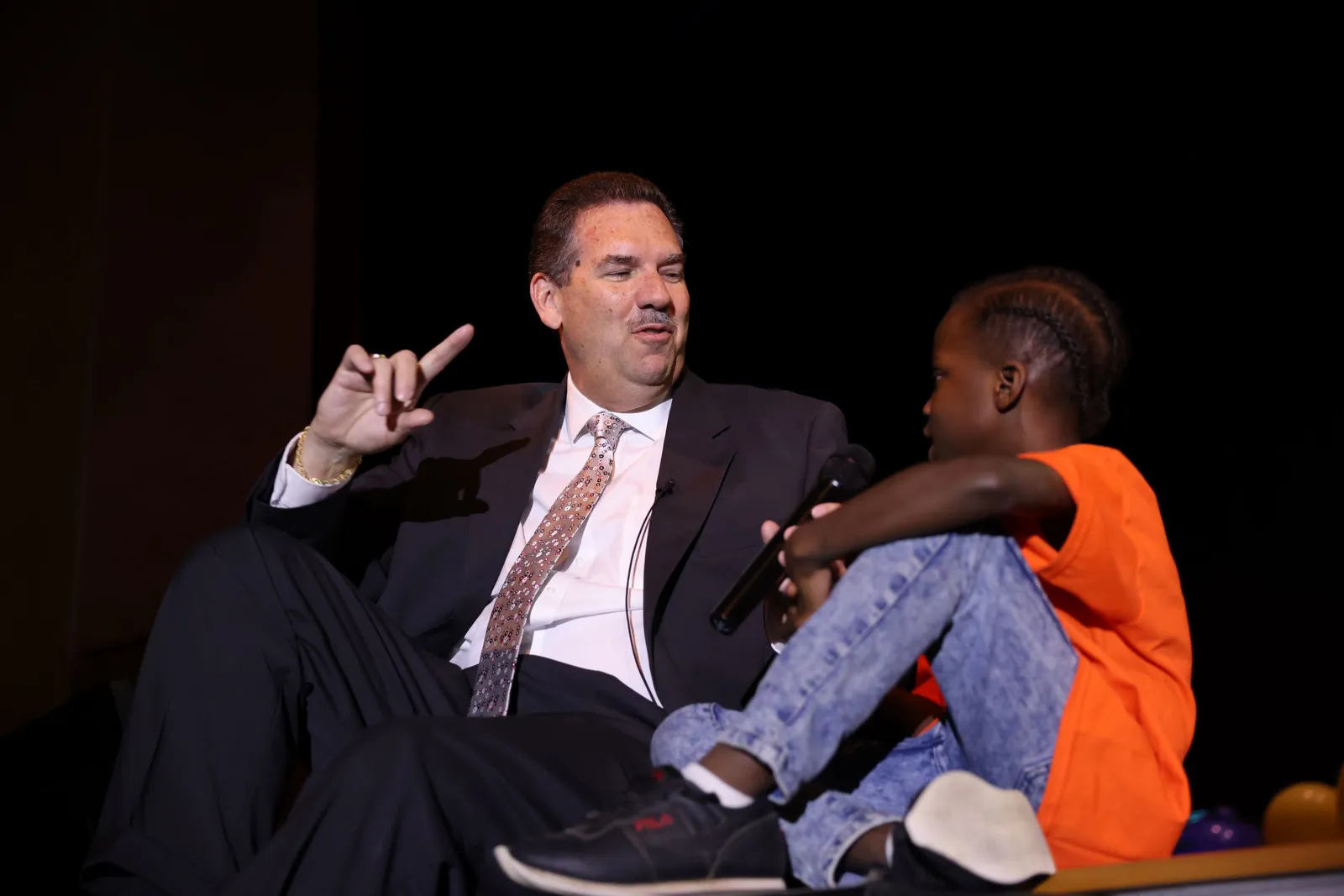 A person wearing a suit and tie sits on a stage next to a young student. The person is holding a microphone in front of the student and speaking.