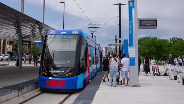 People on train station platform alongside a blue tram with a sign reading "Riverfront."