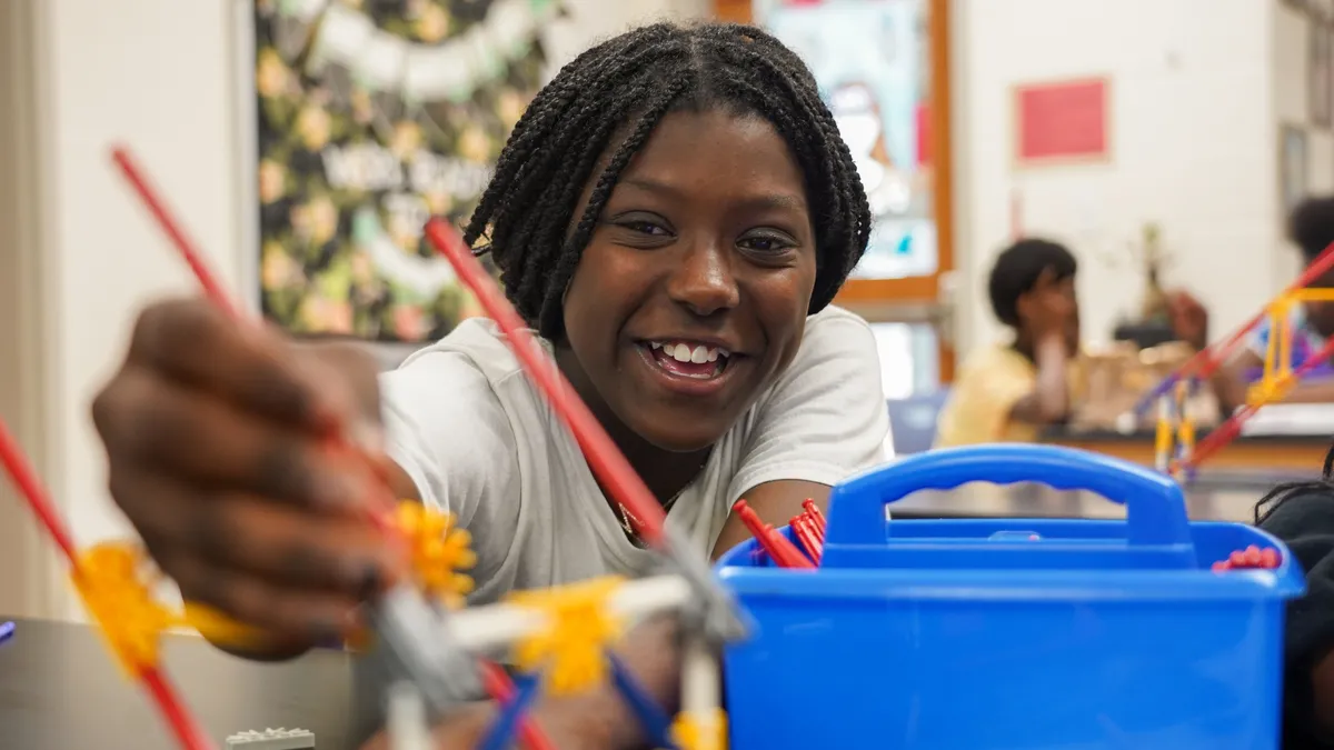 A student sits at a desk, face toward camera, reaches for a paint brush or pen in a blue bin.