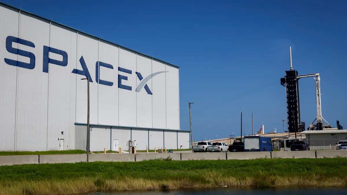 A white SpaceX building on the left and its Falcon 9 rocket on the right on a clear day.