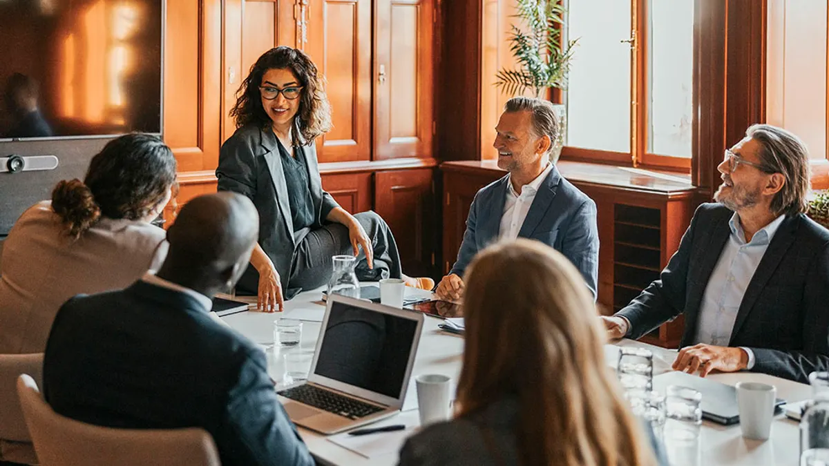 Business team meeting in a wood-paneled boardroom, with a woman presenting while colleagues listen around a table with laptops and documents.