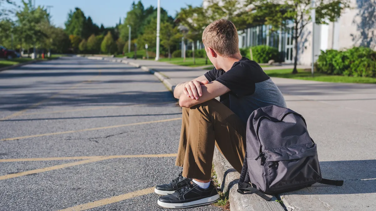 A child sits by the curb of a school with a backpack