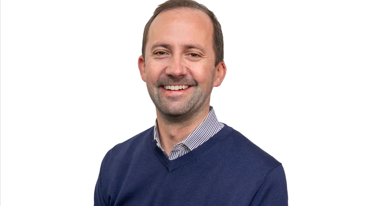 A smiling white man in a blue v-neck sweater in front of a stark white background.