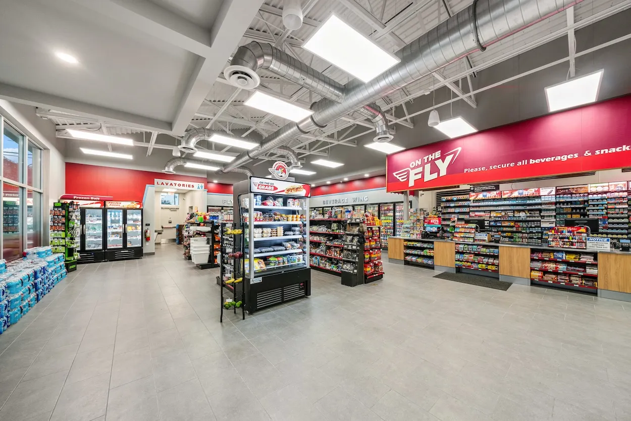 A photo of the interior of a convenience store. there are coolers with drinks and shelves with snacks and other goods. A sign above the checkout area says &quot;On the Fly&quot; while a sign over a doorway to the back reads &quot;lavatories.&quot;