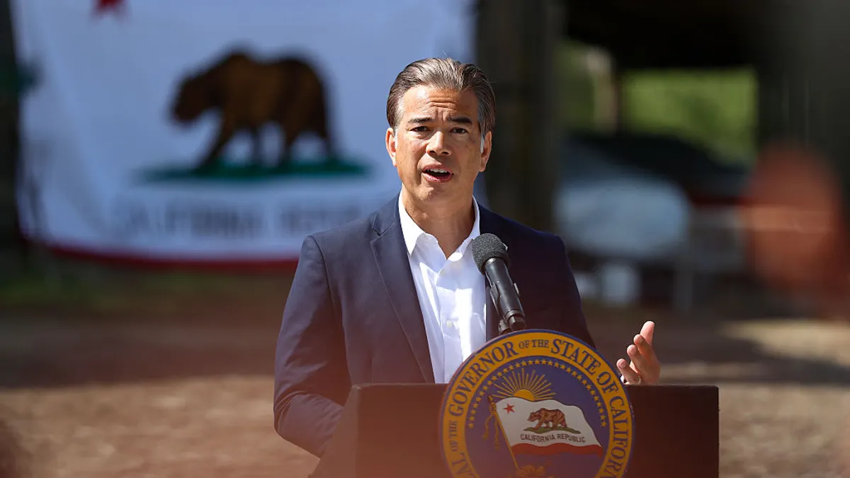 A person stands behind a podium that has a microphone. A state flag of California is seen blurred in the background