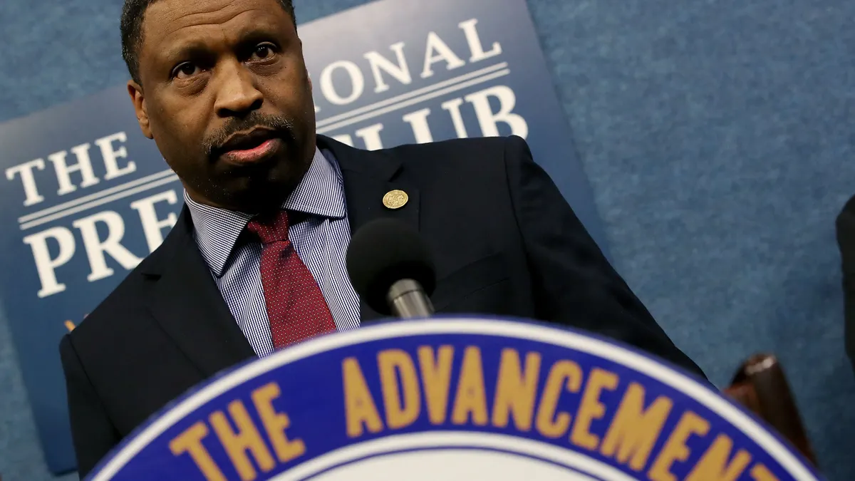 A man in a suit stands at a podium with "The Advancement" visible on the podium.