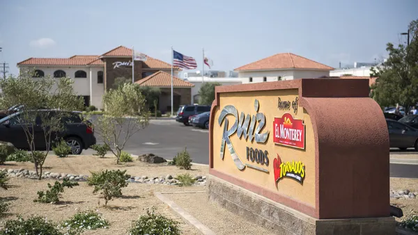 A photo of a stone sign at the edge of a parking lot with a buil;ding in the background. A sign on the building says Ruiz Foods and the stone sign says Home of Ruiz Foods, El Monterey, Tornados.