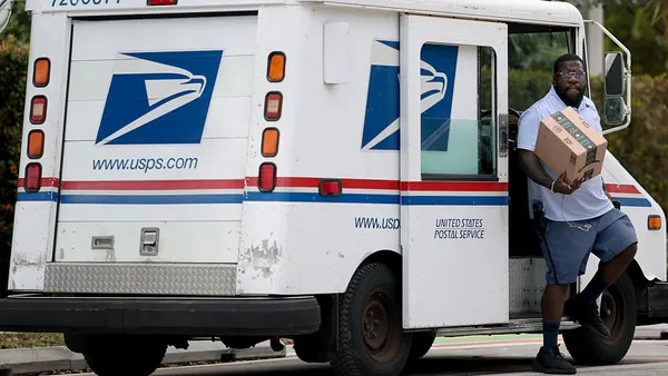 A Postal Service employee steps out of a delivery vehicle with an Amazon-branded package in his hand.