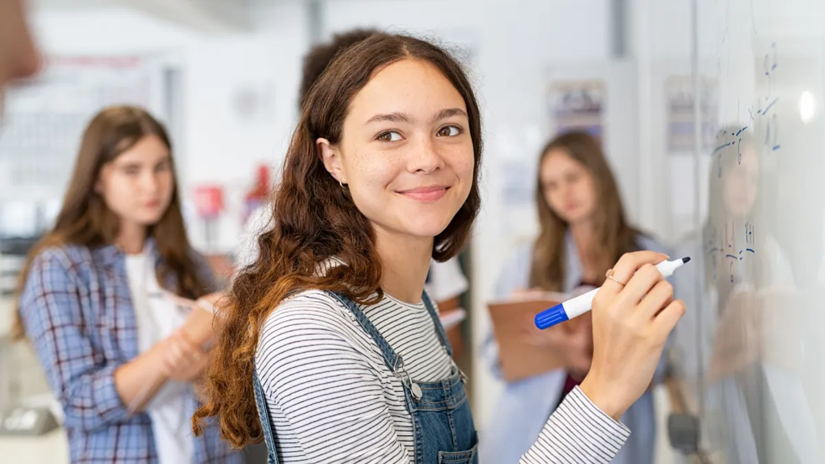 A student writing on a white board and smiling looking over their shoulder