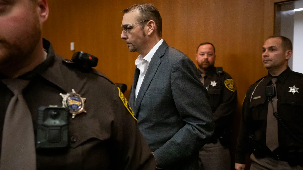 A person is walking into a courtroom. Other people in police uniforms are surrounding the person.