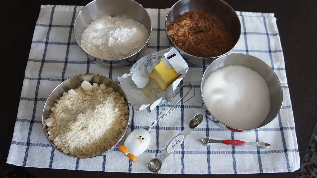 An array of four baking ingredients in silver bowls sitting on a checked kitchen towel with a partial stick of butter, a wire whisk and a measuring spoon sitting near the middle.
