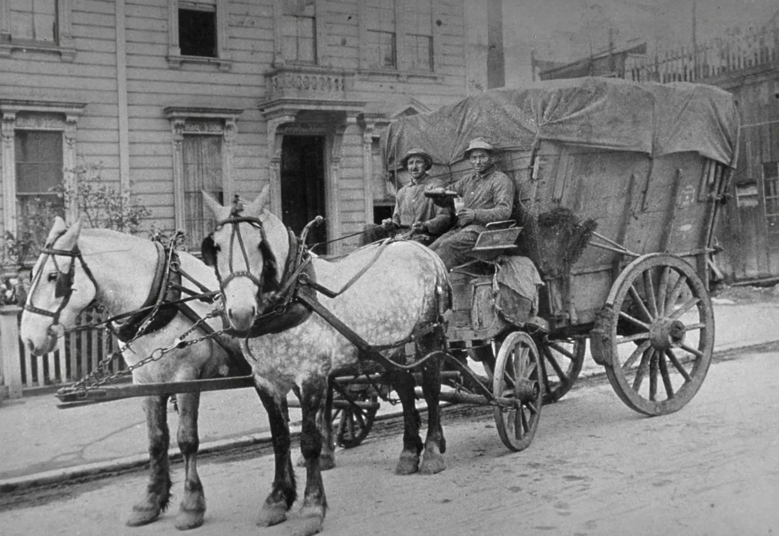 A historic black and white photo fo two men sitting on a horse-drawn carriage used for trash collection