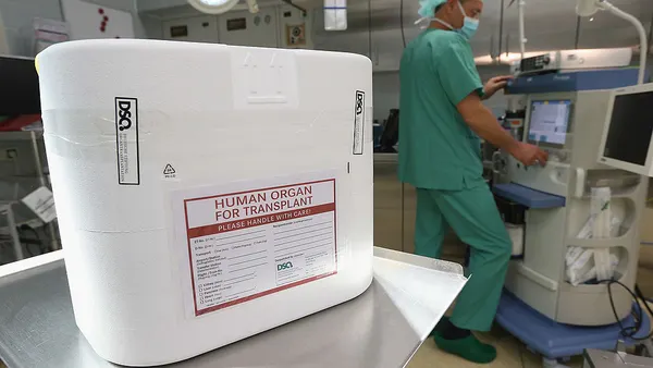 A cooler for an organ transplant sits on a cart in an operating room, as a hospital staffer works behind.