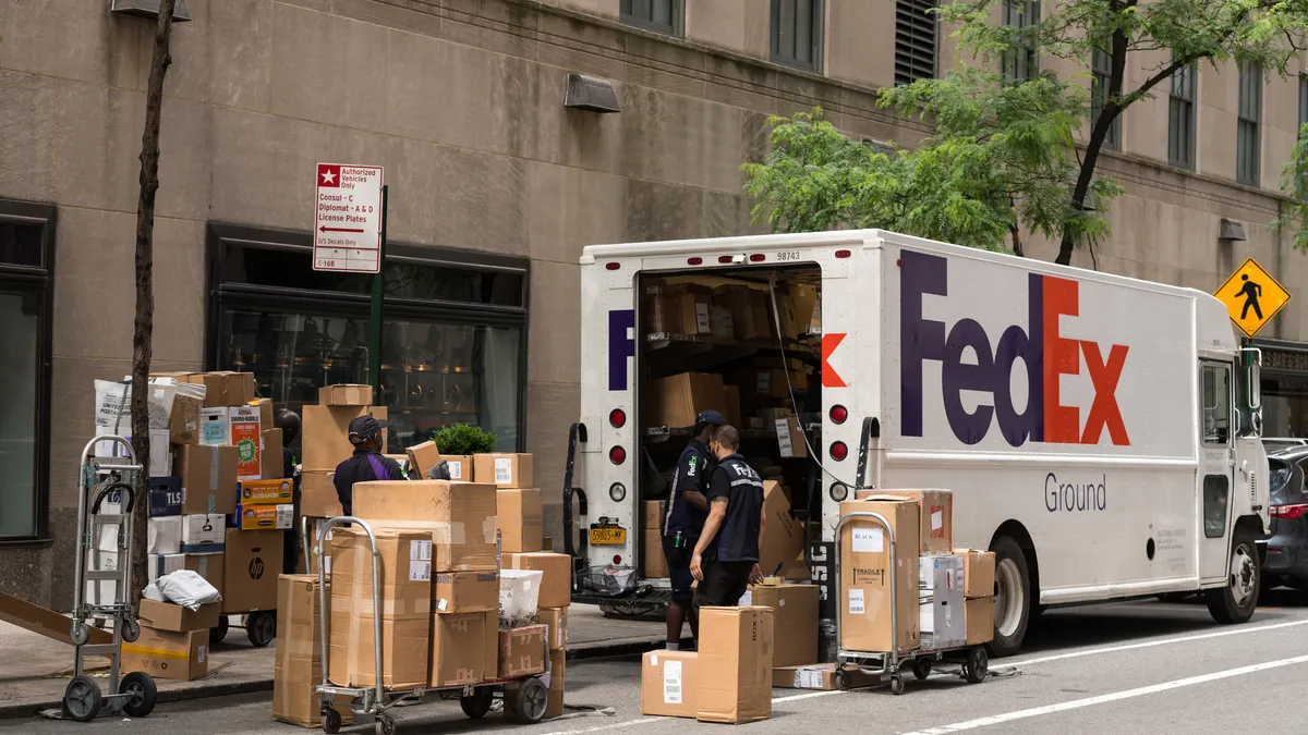 Three people in FedEx uniforms handle packages behind a FedEx delivery vehicle on the street.