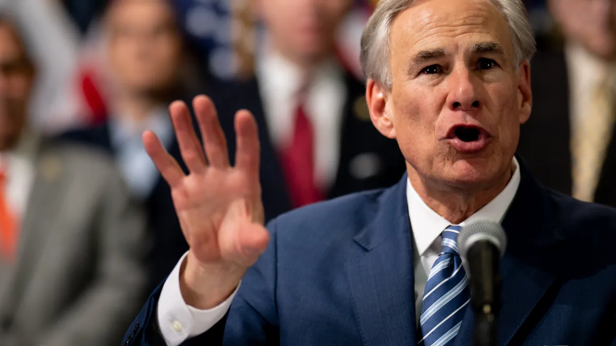 A man gestures before an out-of-focus background of people gathered.