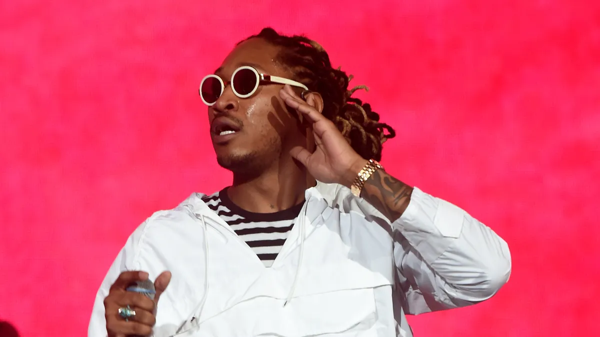 Man with locs, striped black and white shirt, white windbreaker and white sunglasses stands holding a microphone in front of a red background.