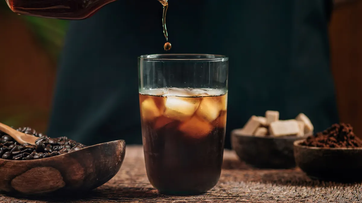 A pitcher pours black coffee into a glass of ice sitting on a brown wooden tablecloth with wooden bowls of different items nearby.