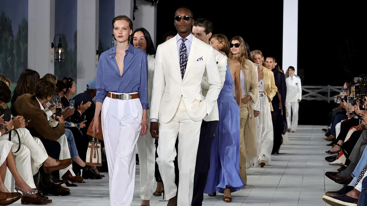 Models in neutral-toned clothing walk down a runway and spectators watch from the sides.