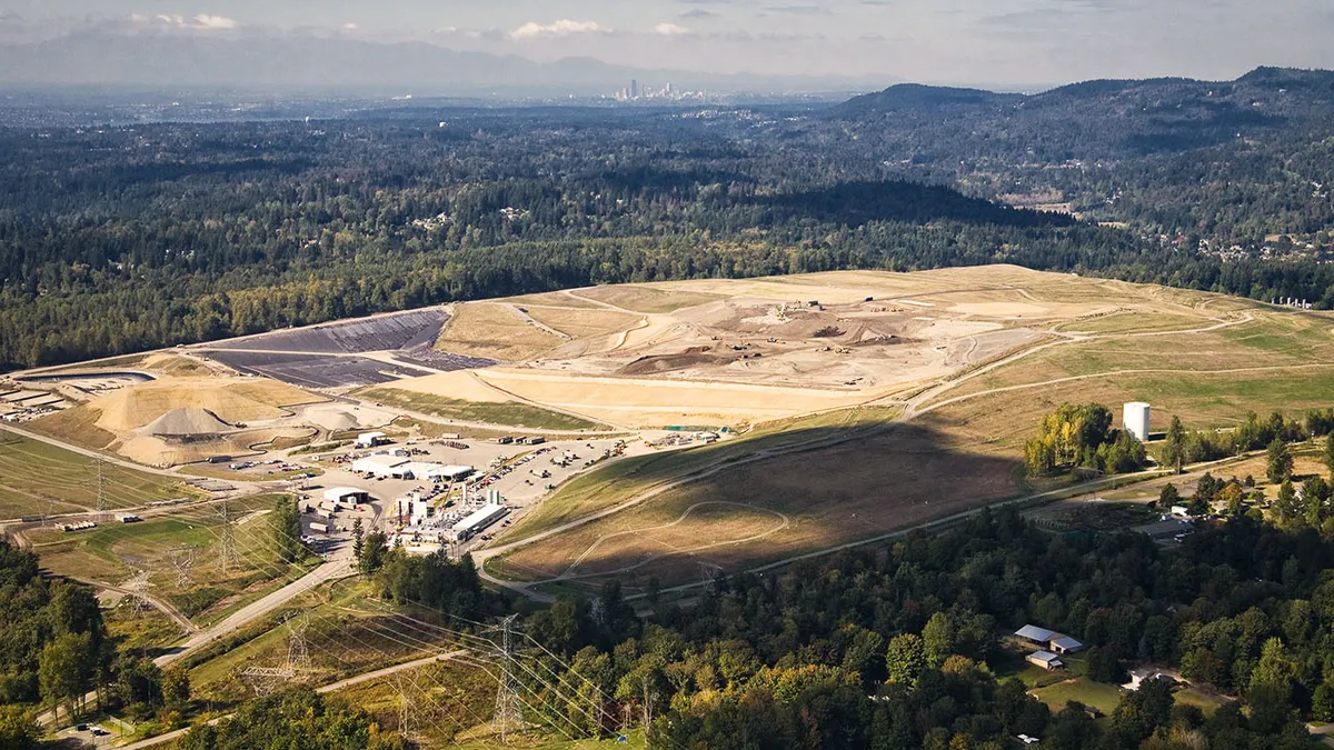 Aerial view of a large landfill near power lines and surrounded by forest. A large city is visible in the distance.